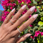 Hand with almond-shaped nails polished in Kitten soft neutral tone showing a smooth manicure outdoors with bright flowers background