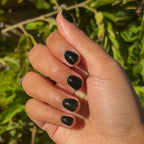 Hand with Latenight ultra-dark olive green quick dry gel polish manicure on natural nails against leafy outdoor background