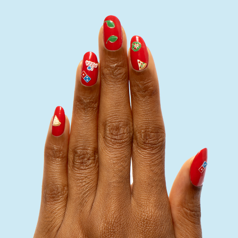 Hand with vibrant red nails featuring Domino’s Pizza Mani Kit quick dry polish and pizza-themed nail art stickers on light blue background