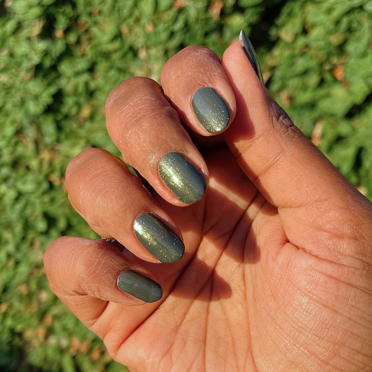 Hand with short nails painted in Martini Chrome showing deep olive green gel polish with golden shimmer finish outdoors