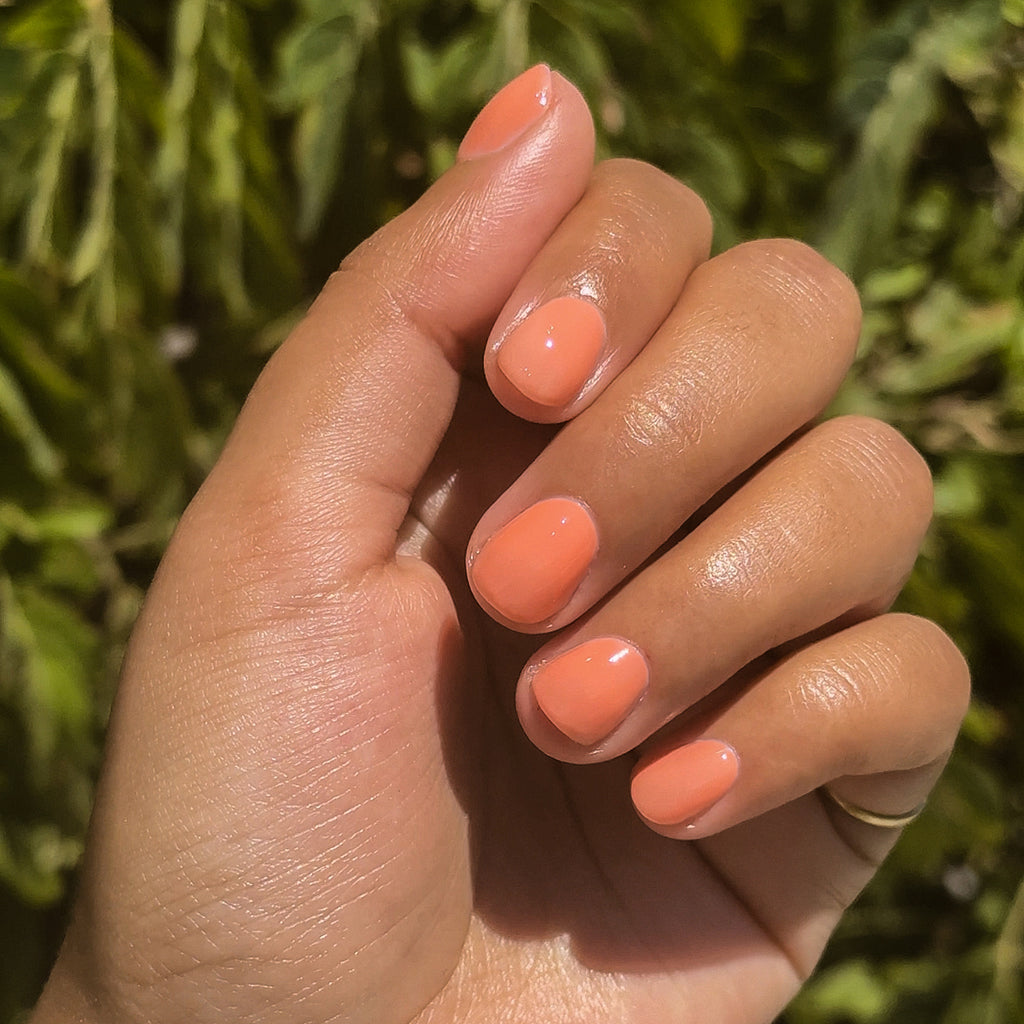 Hand with Peeptoe sheer warm peach quick dry polish on short natural nails against blurred green background