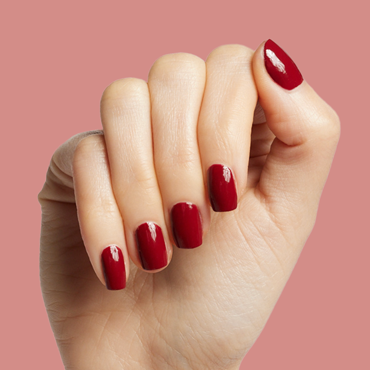 Hand showing glossy deep red manicure with Brick polish against a muted pink background for an at-home mani