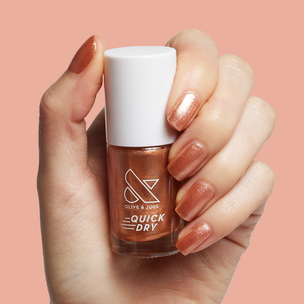Hand holding Penny polish bottle showing shiny copper metallic quick-dry mani on light skin with warm background