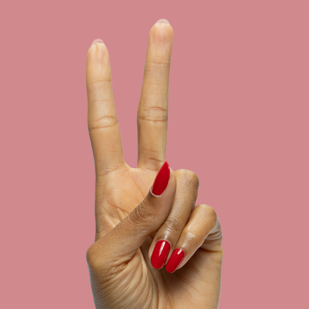 Hand with long oval nails painted in Secret Recipe blue-toned red polish making peace sign on soft pink background