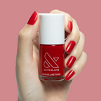 Hand with medium rounded nails in Secret Recipe polish holding Olive and June long-lasting nail polish bottle on pink background