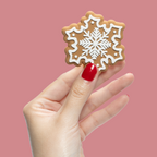 Hand with nails painted in Secret Recipe red polish holding a white-decorated snowflake cookie on pink background