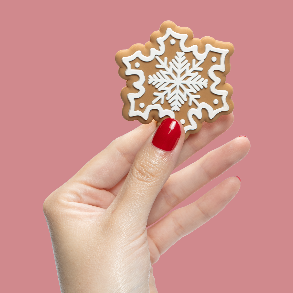 Hand with nails painted in Secret Recipe red polish holding a white-decorated snowflake cookie on pink background