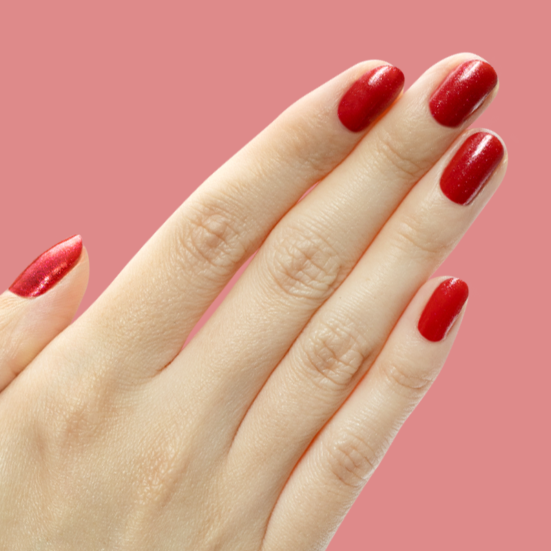 Hand with short nails painted in shimmery berry red gel polish Splash of Cran against a pink background