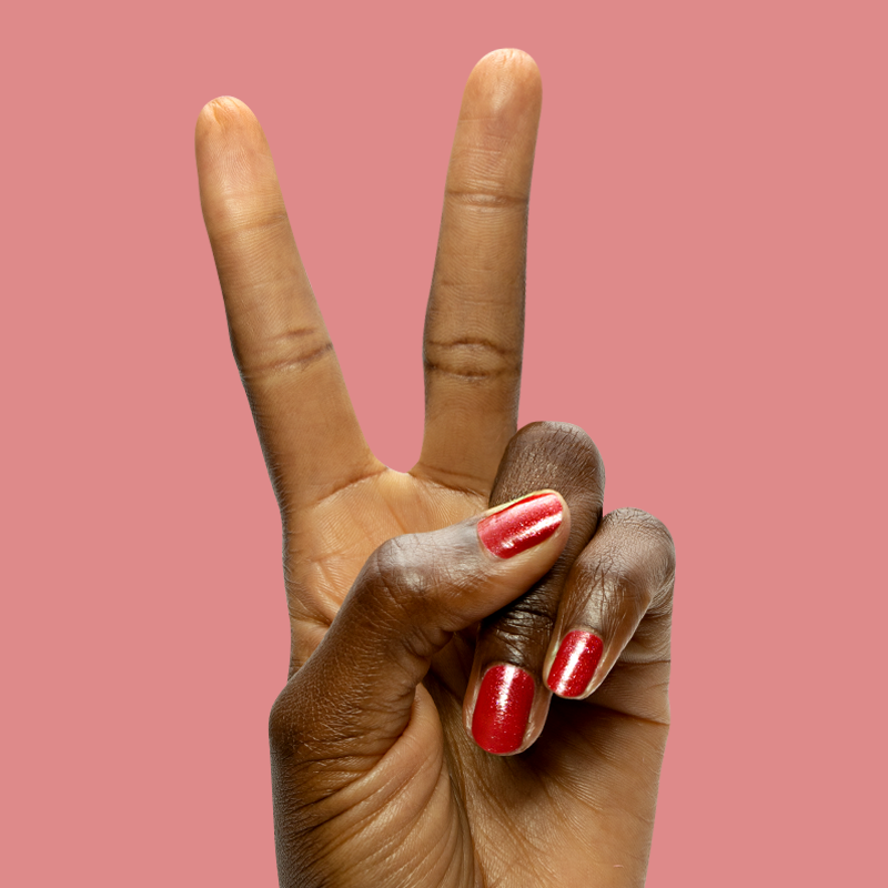 Hand with Splash of Cran gel polish in shimmering pinky-red on short nails against a soft coral background
