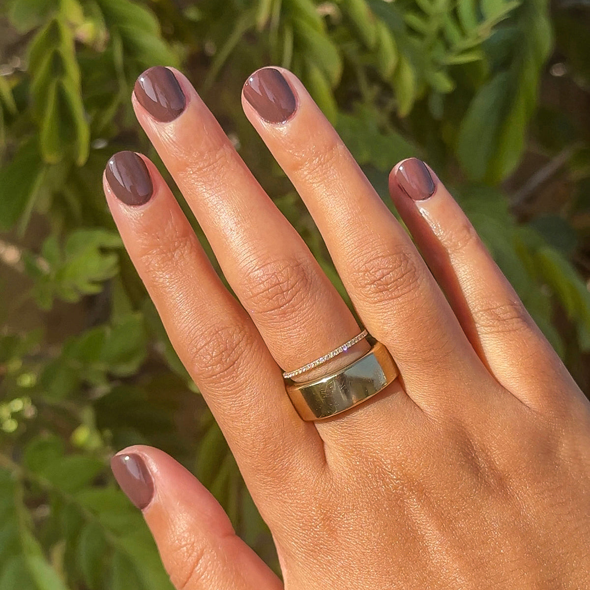 Hand with Spressotini gel polish manicure in rich dark coffee brown on natural short nails with green foliage background
