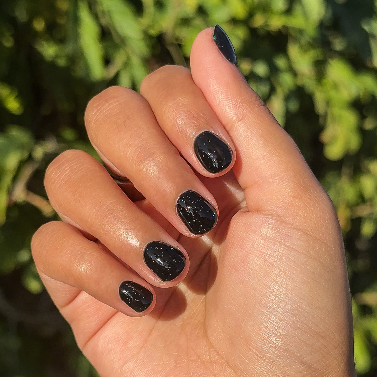 Hand with Stargaze manicure in glossy black polish with delicate rainbow glitter on short natural nails outdoors