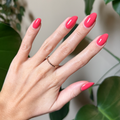 Hand with almond-shaped nails polished in juicy watermelon red using Field Day - Gel against natural leafy background thumbnail