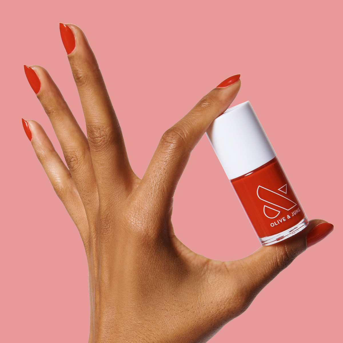 Hand with nails painted bright cherry red holding Olive and June CV polish bottle against pink background