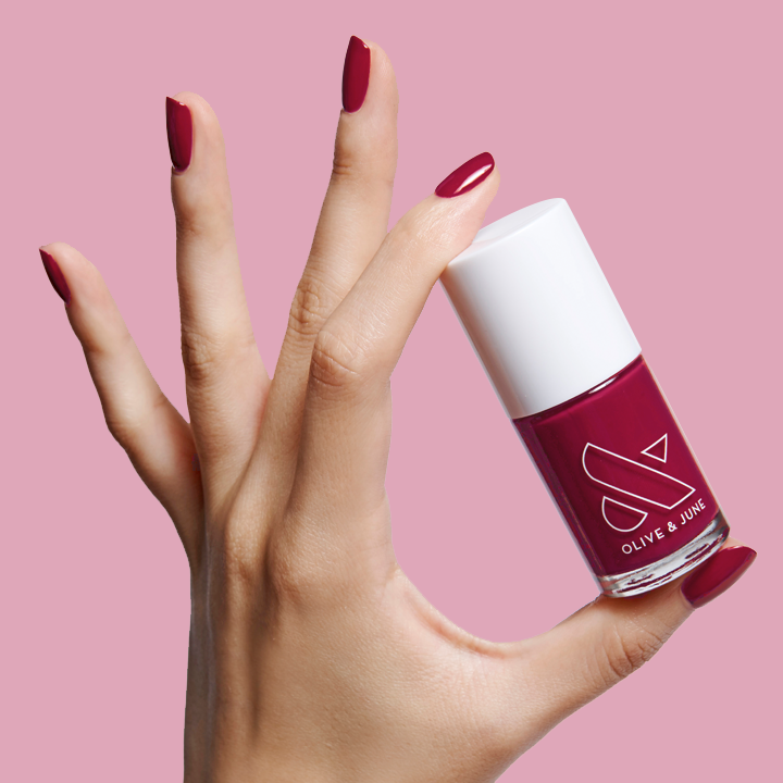 Hand holding a bottle of JG nail polish with nails painted in a rich berry shade against a pale pink background