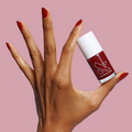 Hand with nails painted in deep bordeaux SC polish holding bottle against a soft pink background