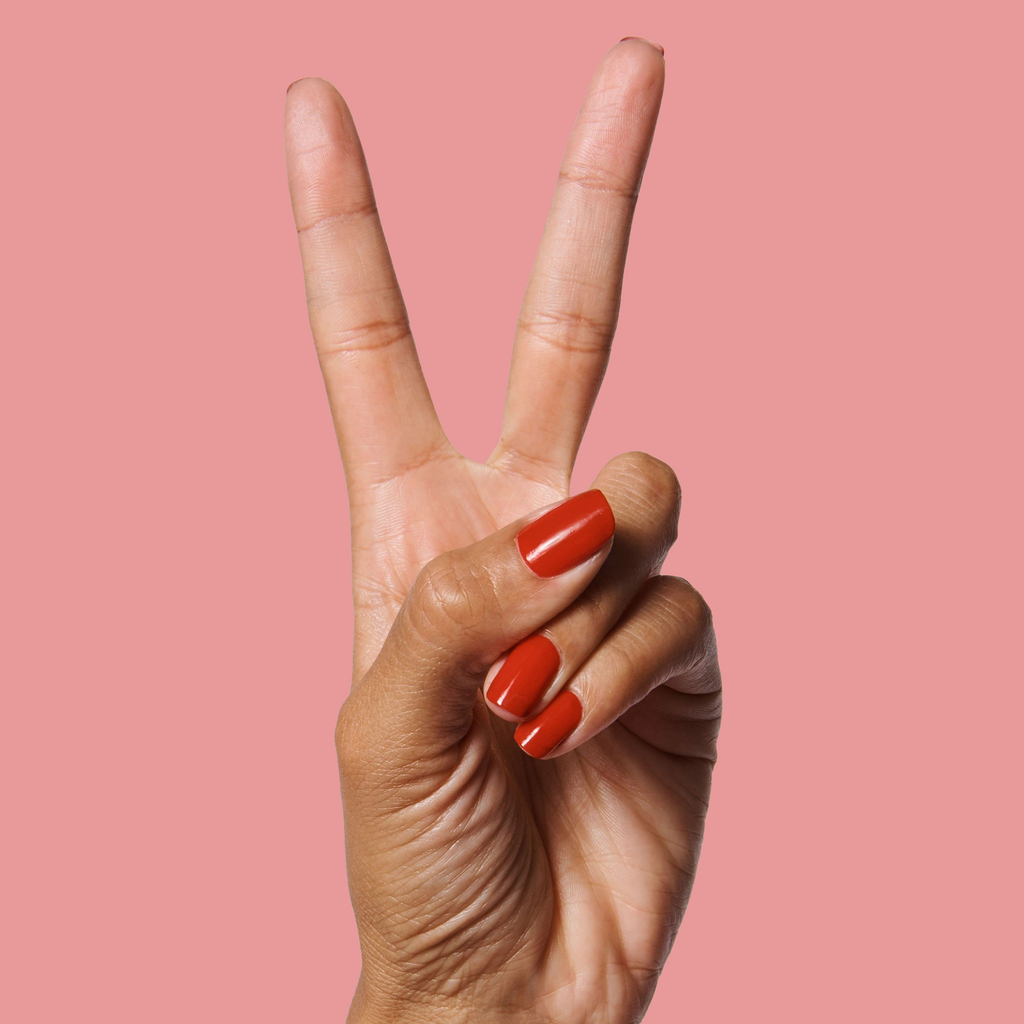 Hand showing nails painted in bright cherry red CV polish by Olive and June against a muted pink background