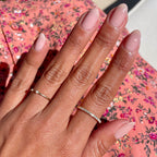 Hand with almond-shaped nails painted in sheer mauve Potpourri gel polish wearing delicate rings on floral fabric background
