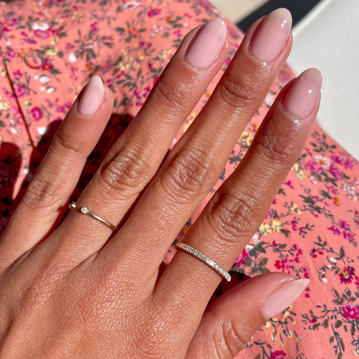 Hand with almond-shaped nails painted in sheer mauve Potpourri gel polish wearing delicate rings on floral fabric background