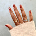 Hand with nails painted in JJ gel polish, a warm cinnamon brown, styled with gold and silver rings and cozy sweater sleeve