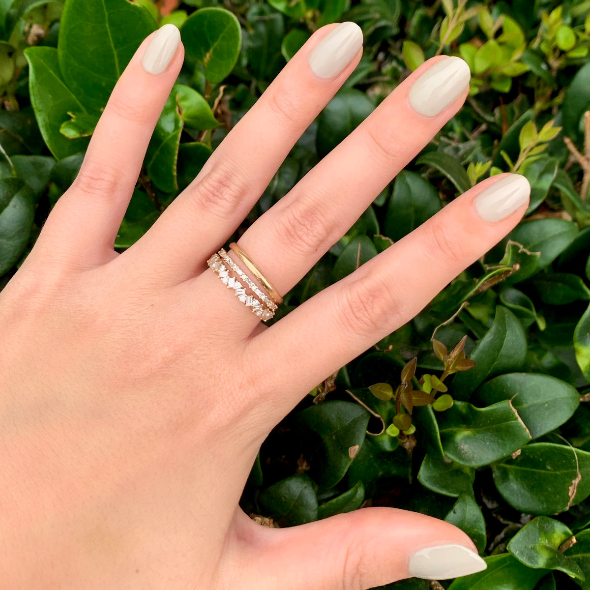 Hand with nails polished in Koala soft grey shade wearing gold rings against green leafy background for a fresh mani