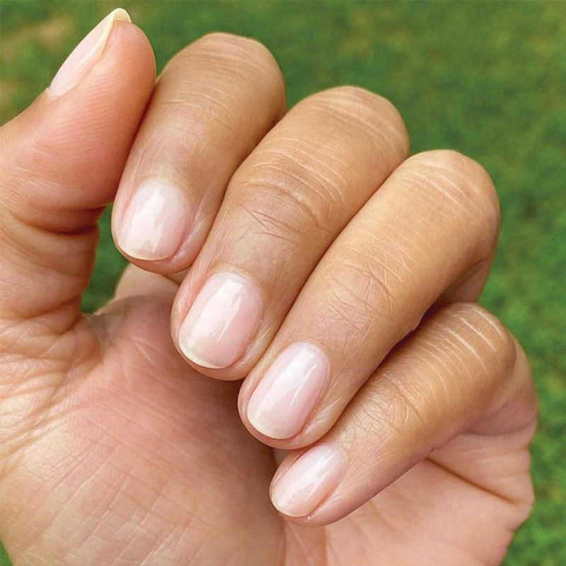 Close-up of a hand with short, natural nails featuring a clear, glossy manicure against a blurred green background