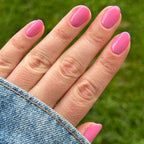 Hand with nails painted in bright pink Taffy gel polish shown outdoors with denim jacket sleeve and green grass background