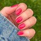 Close-up of hand with Pomodoro quick dry polish in glossy pinky-tomato red on natural nails near denim fabric outdoors