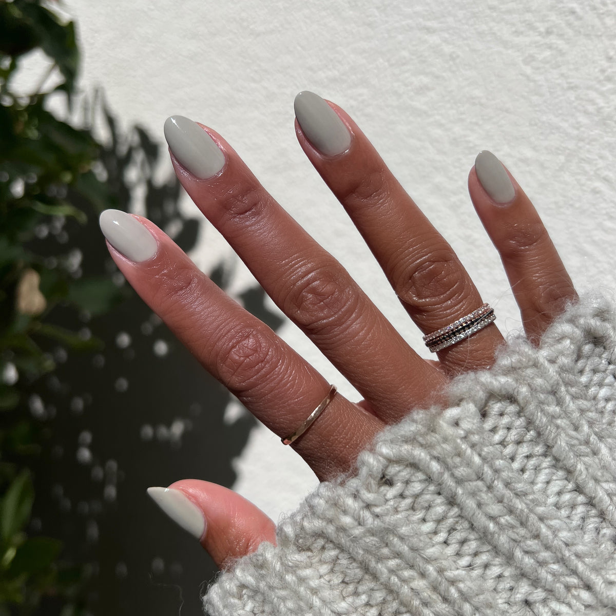 Hand with nails painted in Koala soft grey polish wearing delicate rings and cozy knit sleeve in natural light for a mani
