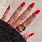 Hand with almond-shaped nails painted in vivid Lollipop quick dry polish wearing bold gold ring on neutral background