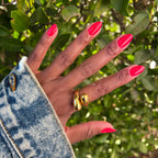 Hand with nails coated in vibrant orangey red Picante polish shown outdoors beside green leaves for manicure display