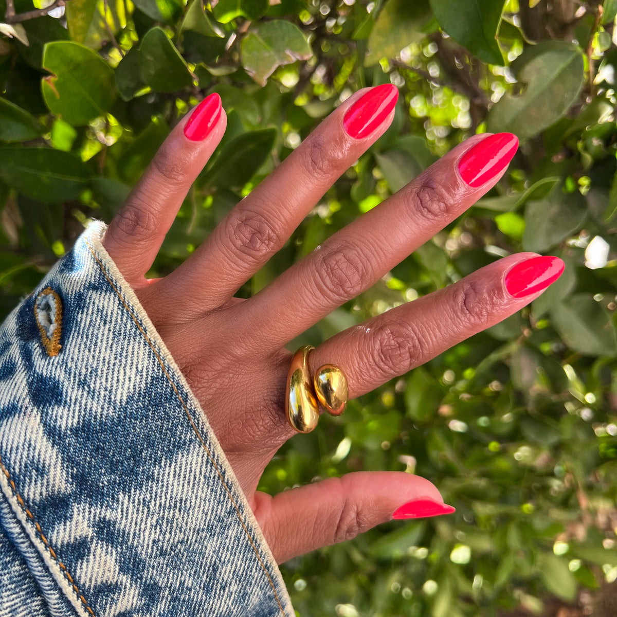 Hand with nails coated in vibrant orangey red Picante polish shown outdoors beside green leaves for manicure display