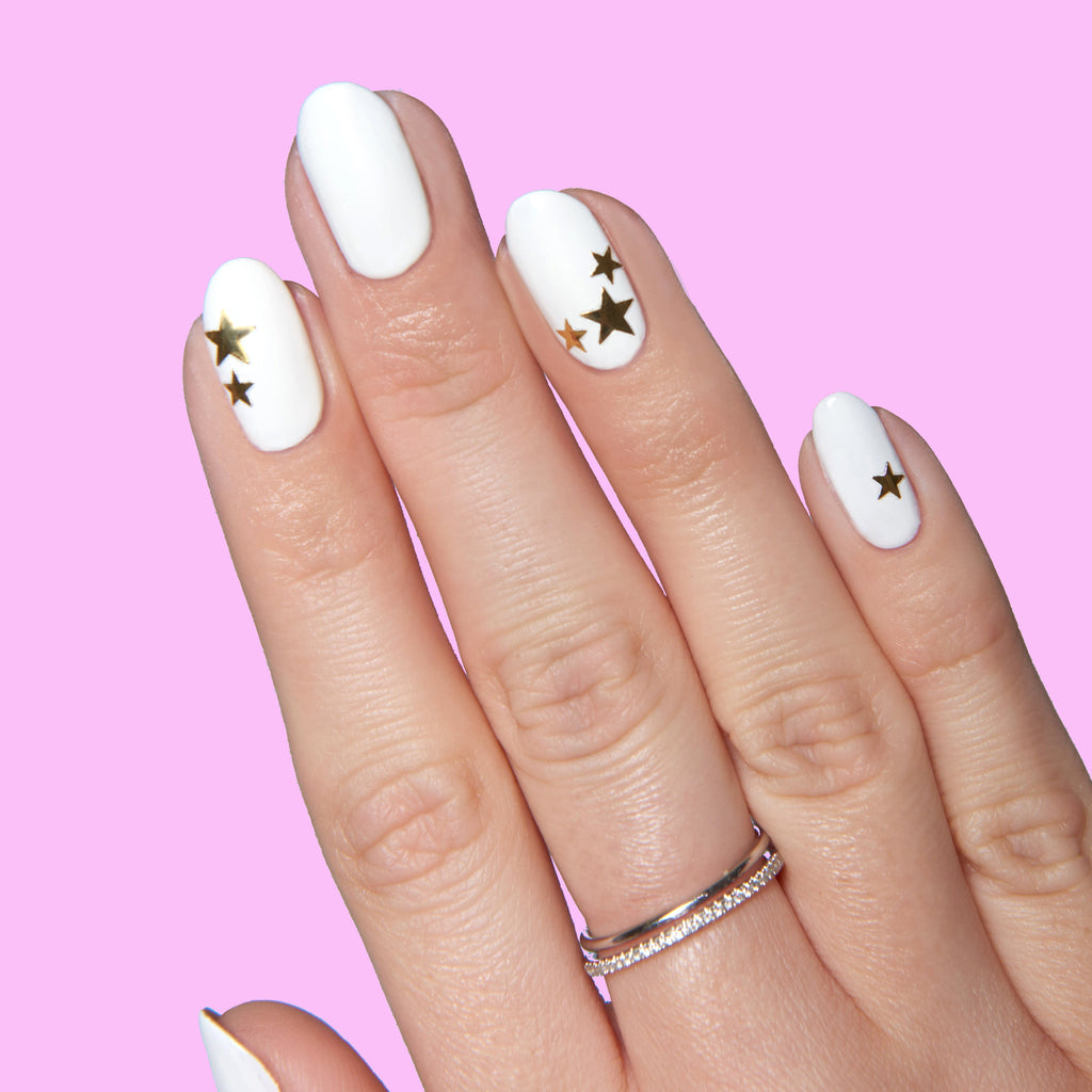 Hand with white polish and gold Stars nail stickers arranged on several nails for Stars manicure on pink background