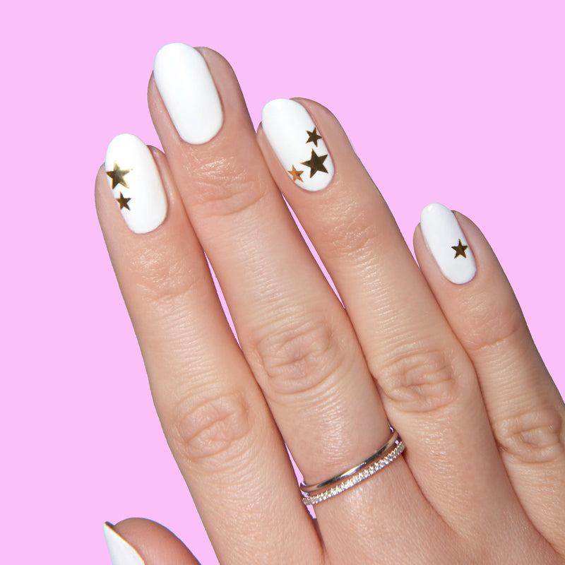 Hand with white polish and gold Stars nail stickers arranged on several nails for Stars manicure on pink background
