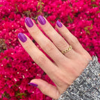 Hand with nails painted bright neon purple Grape Soda polish showing smooth glossy manicure with vibrant floral background