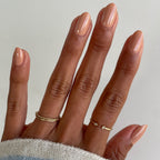 Fingers with nails polished in Howdy neutral beige showing a glossy manicure and gold rings on a light background
