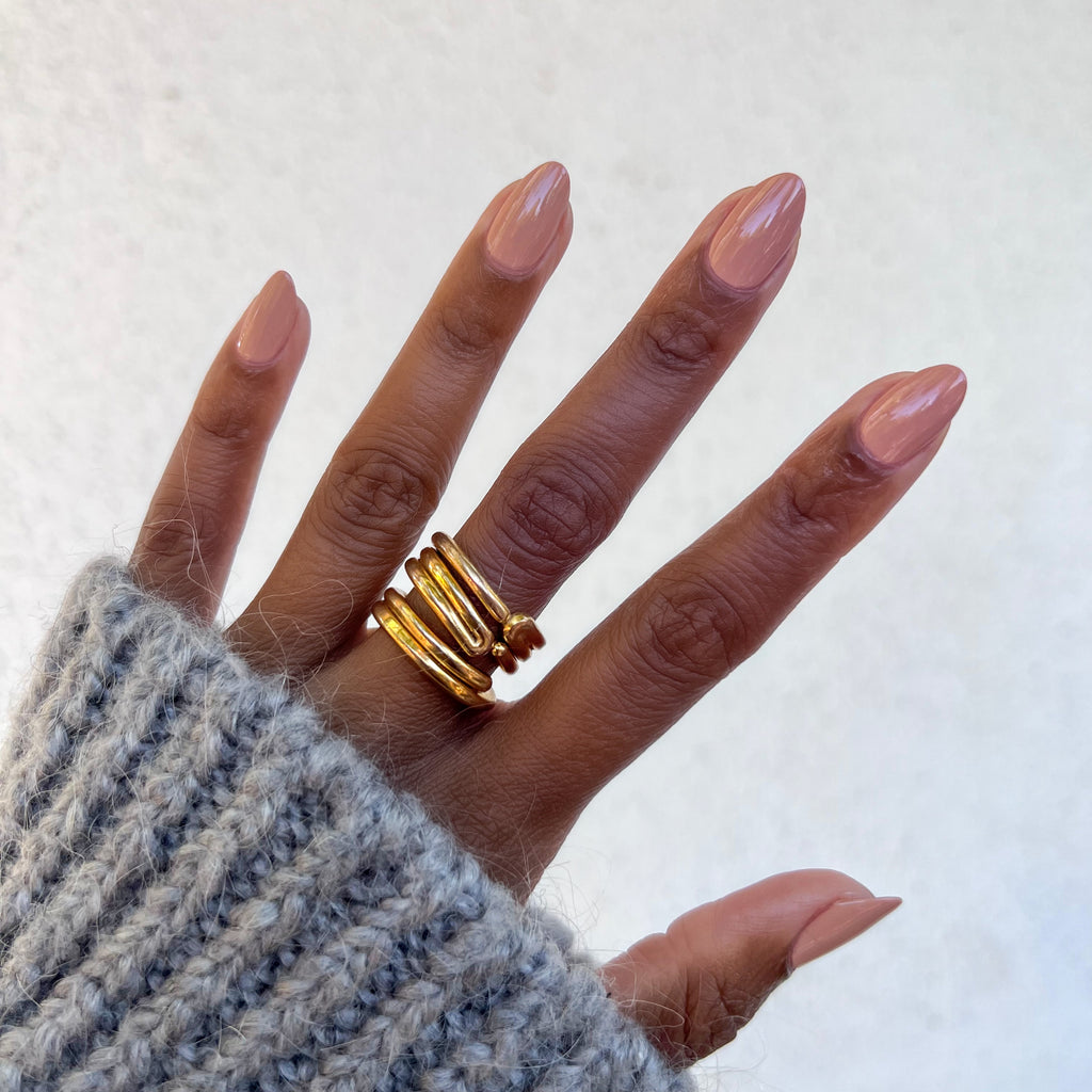 Hand showing nails polished in Howdy neutral beige with terracotta undertones and gold rings against a white background