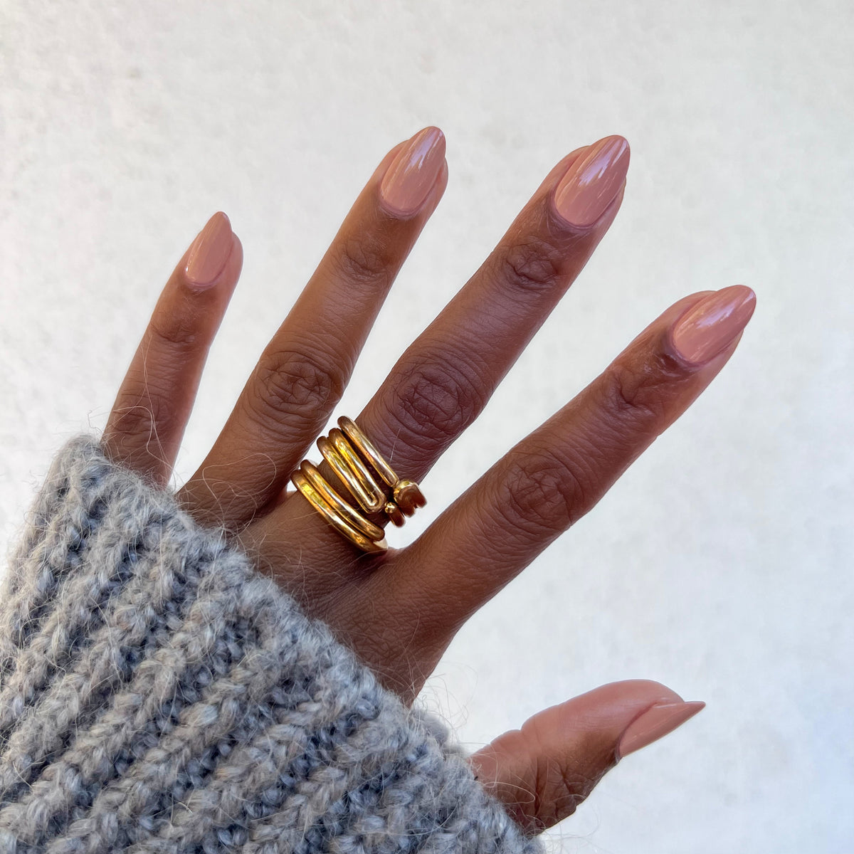 Hand showing nails polished in Howdy neutral beige with terracotta undertones and gold rings against a white background