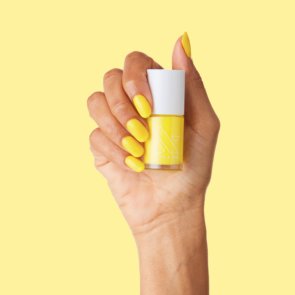 Hand holding bottle of Lemony Lemon yellow nail polish with matching bright mani against yellow background