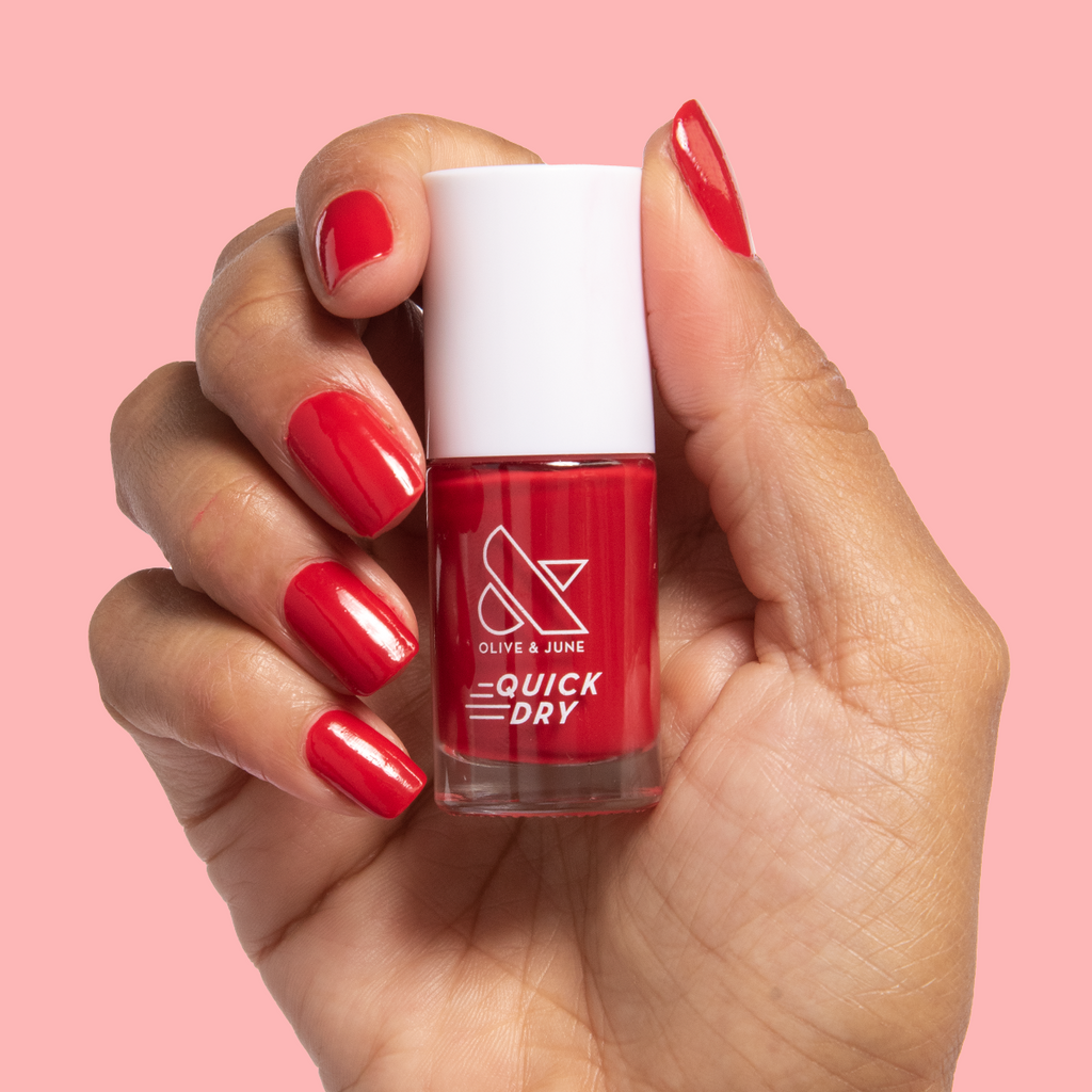 Hand holding a bottle of Smooch quick dry nail polish with matching red painted nails on pink background